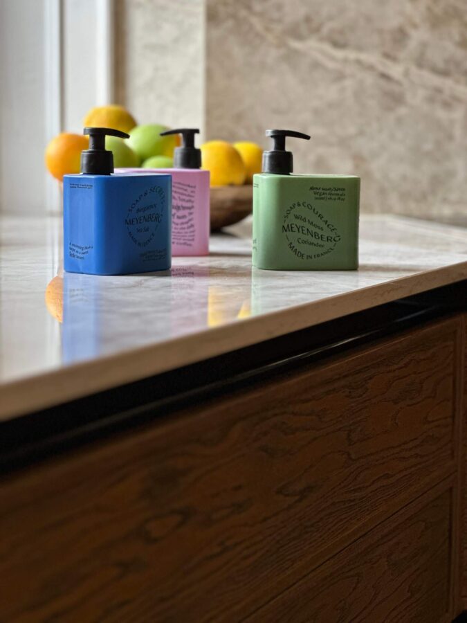 Tre Meyenberg handtvålar i grönt, blått och lila på köksbänk med fruktskål i bakgrunden.