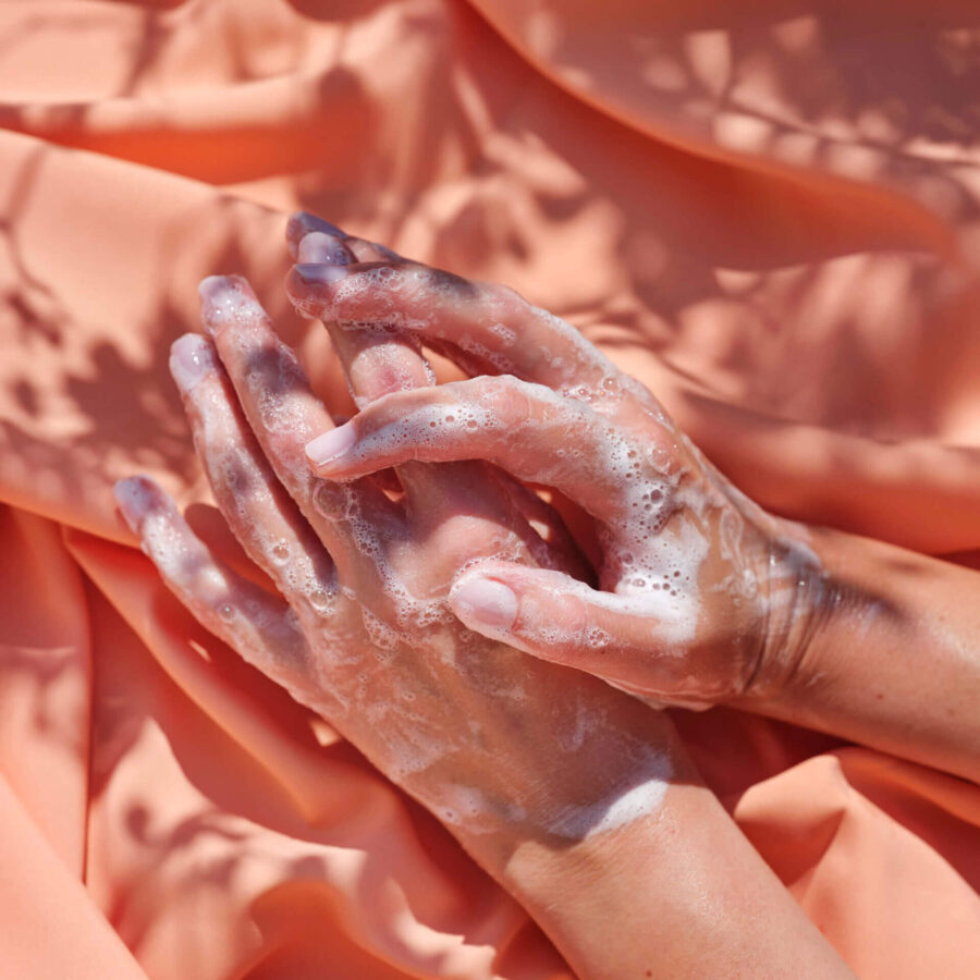 Händer täckta av tvålskum framför persikofärgat tyg, visar produktens rengörande effekt.
