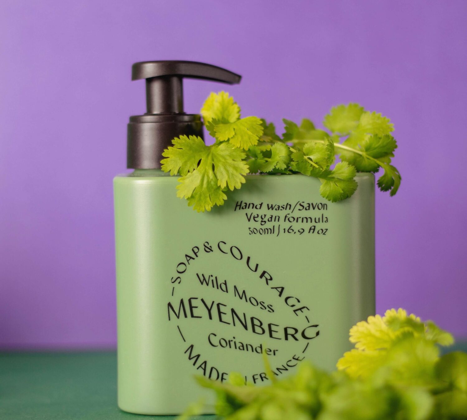 Meyenbergs gröna handtvål i doften courage. Inramad av koriander mot en grön lila bakgrund.