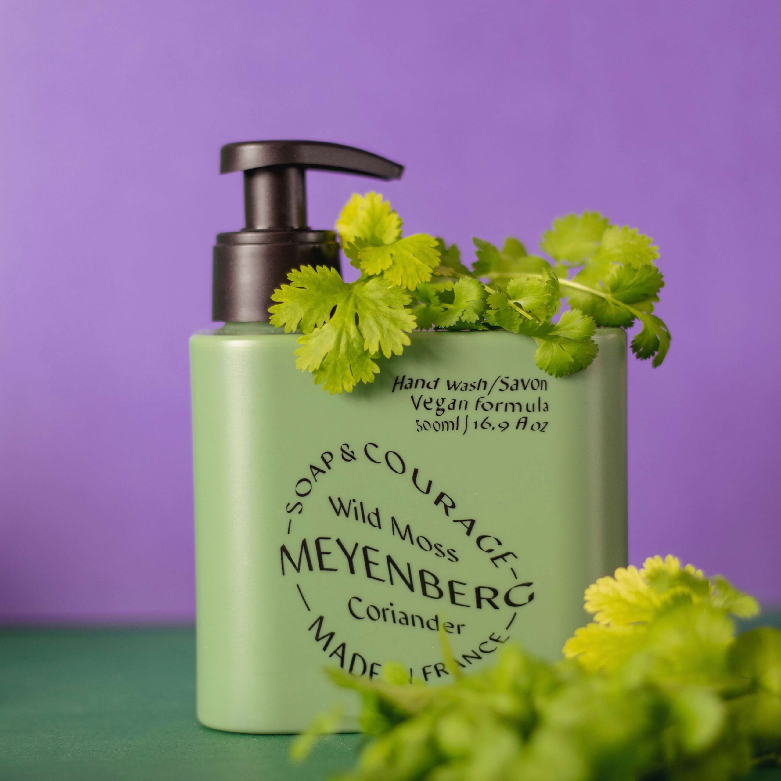 Meyenbergs gröna handtvål i doften courage. Inramad av koriander mot en grön lila bakgrund.